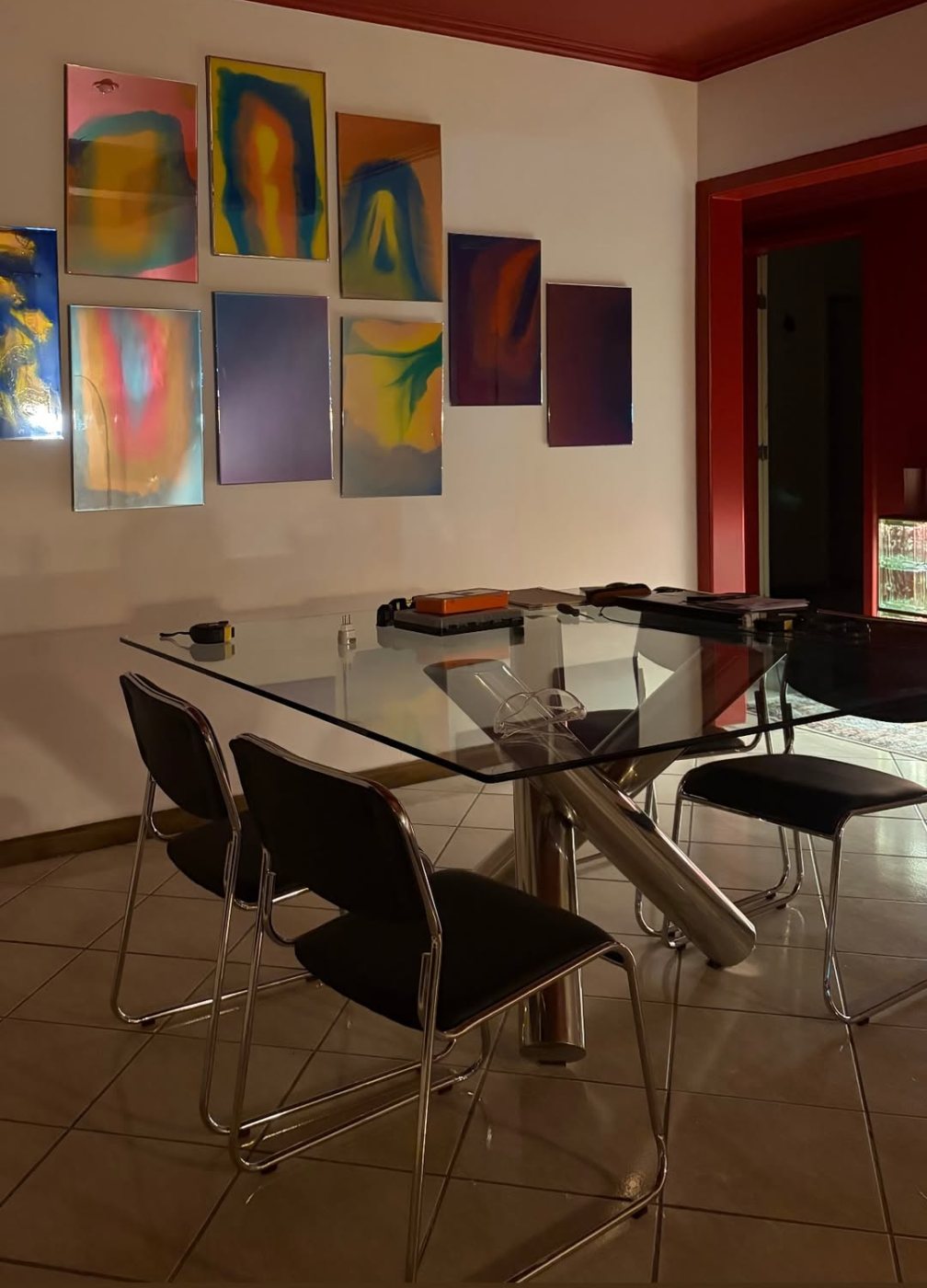 Parede do ateliê com vários espelhos coloridos pendurados, acima de uma mesa de vidro, sob um teto vermelho.